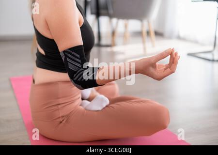 Frau, die im Kreuz auf Yogamatte sitzt, in Lotusposition, mit Sportbekleidung, die sich auf das Atmen in einer modernen Wohnung konzentriert Stockfoto