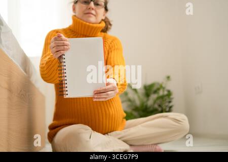 Schwangere Frau, die ein leeres Notizbuch hält. Konzentrieren Sie sich auf die leere Seite im Vordergrund. Sie sitzt drinnen und trägt einen gelben Strickpullover, was eine ruhige und gemütliche Atmosphäre schafft. Stockfoto