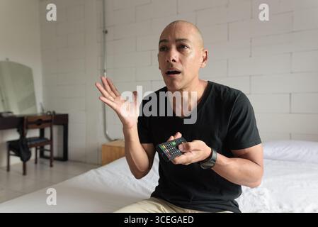 Mann mittleren Alters in schwarzem T-Shirt, der auf dem Bett sitzt und Fernsehen in einem minimalistischen weißen Apartment guckt, das schockiert und überrascht aussieht. Freizeitleben Stockfoto