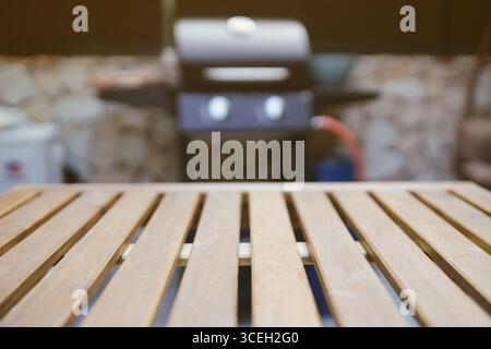 Nahaufnahme eines hölzernen Außentisches mit einem verschwommenen Gasgrill im Hintergrund. Backyard BBQ-Einrichtung bereit für Sommerkochen und gesellige Zusammenkünfte. Stockfoto