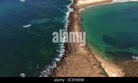 Aus der Vogelperspektive auf einen Wellenbrecher, der den dunkelblauen Ozean vom helleren türkisfarbenen Wasser der Bucht trennt und einen auffälligen Kontrast von Farbe und Textur schafft, Newcastle, NSW, Australien. Stockfoto