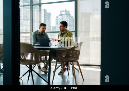 Zwei junge Männer arbeiten zusammen an einem Projekt, sitzen mit einem Laptop an einem Tisch. Die Einrichtung ist ein offener Bürobereich mit Glasfenstern und Blick auf einen Stockfoto