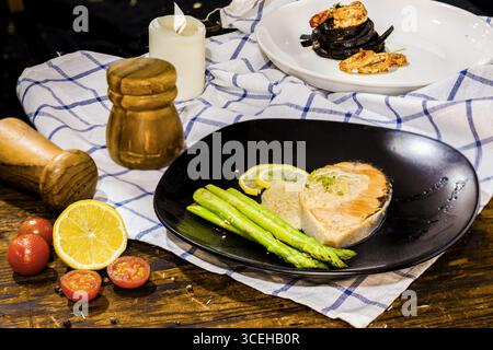 Lachssteak mit grünem Spargel; Schwarze Fettuccine-Spaghetti-Pasta mit Steingarnelen und Tintenfisch-Belag mit grüner Petersilie auf Holztisch, Serviette. Stockfoto