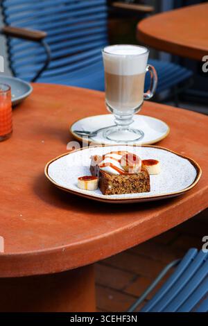 Schaumiger Latte auf einem hohen Glas und eine Scheibe schön präsentiertes Bananenbrot mit Bananenscheiben und Karamellsauce. Im Freien, Terrassenlandschaft, lokaler Brunch Stockfoto