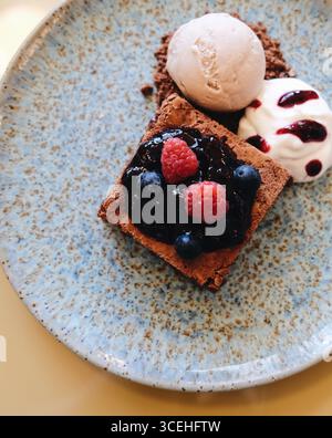 Schön präsentiertes Dessert im Restaurant: Schokoladenbrownie mit Beerenmarmelade und frischen Beeren, Eisschaufel und Schlagsahne Stockfoto