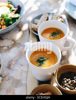 Zwei weiße Tassen warmer, cremiger Butternusskürbis- oder Kürbissuppe, garniert mit frischen Kräutern, serviert auf einem Holzbrett mit Brot und Samen auf einer sonnigen Sonne Stockfoto