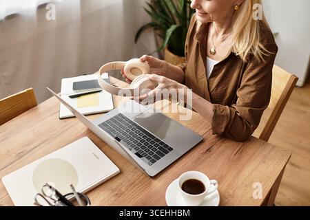 Schöne Seniorin sitzt an einem Holztisch und hält Kopfhörer, während sie an ihrem Laptop arbeitet. Stockfoto