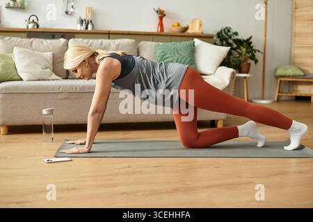 Schöne Seniorin übt tagsüber Yoga auf einer Matte in einem gemütlichen Wohnzimmer. Stockfoto