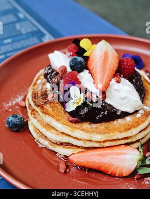 Wunderschön präsentierte amerikanische Pfannkuchen mit Sirup, frischen Beeren, Blumen und Creme auf einem eleganten Dessertteller in einem Café-Stil Stockfoto
