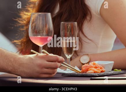 Nahaufnahme eines Paares, das Sushi mit Essstäbchen und Gläsern Wein auf dem Tisch im Freien genießt, romantischer Essensmoment. Stockfoto