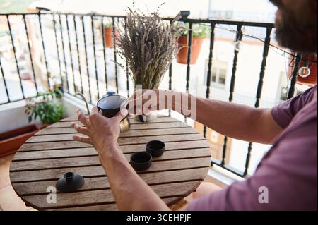 Ein Mann genießt Tee auf einem gemütlichen Balkon mit Holzmöbeln und Topfpflanzen. Die Umgebung ruft Ruhe hervor, was Momente der Entspannung und der Entspannung suggeriert Stockfoto