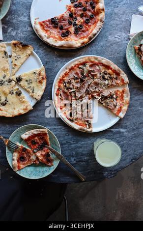 Blick von oben auf italienische Pizzen auf einem rustikalen Tisch, einschließlich Sorten mit Oliven, Pilzen, Käse und Schinken. Serviert mit Getränken Stockfoto