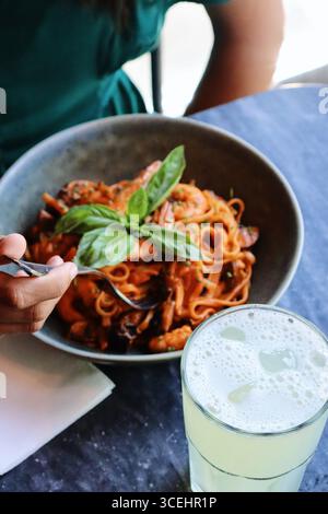 Teller mit Tomatensauce, garniert mit frischem Basilikum und einem Limonadengetränk, Mädchen im Hintergrund hält eine Gabel mit hinterem, dunklem Hintergrund Stockfoto