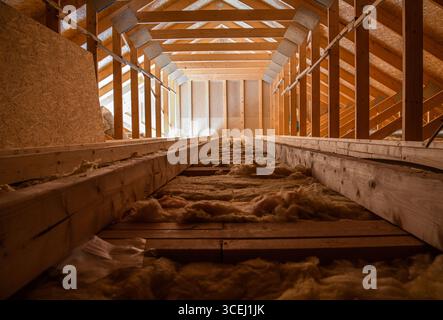Sonnenlicht filtert durch die Dachsparren eines Dachgeschosses, beleuchtet Holzbalken und weiche Isolierung, die über den Boden verteilt sind, und schafft eine warme Atmosphäre Stockfoto