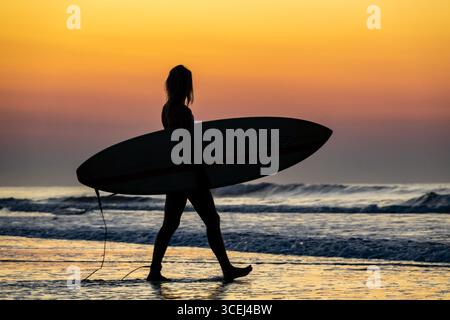 Isle Of Palms, Usa. August 2025. Eine Surferin, die von Sonnenaufgang umrahmt wird, wandert zum Wasser, um die frühen Wellen am Front Beach, 18. August 2025 in Isle of Palms, South Carolina, zu fangen. Hohe Wellen und potenzieller Sturm serge wird später in der Woche erwartet, als Hurrikan Erin, ein Sturm der Kategorie 4, vor der Küste vorbeizieht. Quelle: Richard Ellis/Richard Ellis/Alamy Live News Stockfoto
