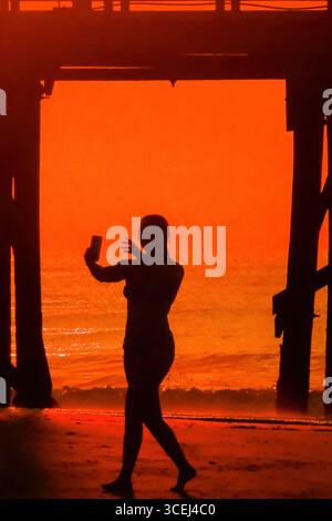 Isle Of Palms, Usa. Januar 2016. Eine junge Frau, die beim Sonnenaufgang über dem Atlantischen Ozean eine Silhouettierung zeigt, nimmt ein Telefonvideo von Under Front Beach Fishing Pier auf der Isle of Palms, South Carolina, am 18. August 2025 auf. Hohe Wellen und potenzieller Sturm serge wird später in der Woche erwartet, als Hurrikan Erin, ein Sturm der Kategorie 4, vor der Küste vorbeizieht. Quelle: Richard Ellis/Richard Ellis/Alamy Live News Stockfoto