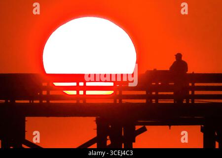 Isle Of Palms, Usa. Januar 2016. Ein Fischer, der beim Sonnenaufgang über dem Atlantik auf dem Front Beach Fishing Pier, 18. August 2025 in Isle of Palms, South Carolina, eine Silhouette bildet. Hohe Wellen und potenzieller Sturm serge wird später in der Woche erwartet, als Hurrikan Erin, ein Sturm der Kategorie 4, vor der Küste vorbeizieht. Quelle: Richard Ellis/Richard Ellis/Alamy Live News Stockfoto