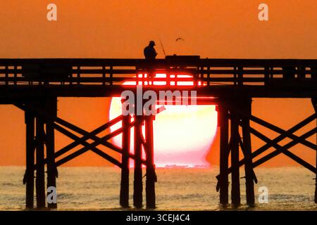 Isle Of Palms, Usa. Januar 2016. Ein Fischer, der beim Sonnenaufgang über dem Atlantik auf dem Front Beach Fishing Pier, 18. August 2025 in Isle of Palms, South Carolina, eine Silhouette bildet. Hohe Wellen und potenzieller Sturm serge wird später in der Woche erwartet, als Hurrikan Erin, ein Sturm der Kategorie 4, vor der Küste vorbeizieht. Quelle: Richard Ellis/Richard Ellis/Alamy Live News Stockfoto