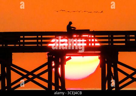 Isle Of Palms, Usa. Januar 2016. Ein Fischer, der beim Sonnenaufgang über dem Atlantik auf dem Front Beach Fishing Pier, 18. August 2025 in Isle of Palms, South Carolina, eine Silhouette bildet. Hohe Wellen und potenzieller Sturm serge wird später in der Woche erwartet, als Hurrikan Erin, ein Sturm der Kategorie 4, vor der Küste vorbeizieht. Quelle: Richard Ellis/Richard Ellis/Alamy Live News Stockfoto