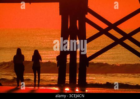 Isle Of Palms, Usa. Januar 2016. Junge Frauen, die beim Sonnenaufgang über dem Atlantischen Ozean eine Silhouettenansicht haben, beobachten den Angelpier Under Front Beach am 18. August 2025 in Isle of Palms, South Carolina. Hohe Wellen und potenzieller Sturm serge wird später in der Woche erwartet, als Hurrikan Erin, ein Sturm der Kategorie 4, vor der Küste vorbeizieht. Quelle: Richard Ellis/Richard Ellis/Alamy Live News Stockfoto