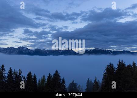 Atemberaubender Blick auf die schneebedeckten Berggipfel, die in der Abenddämmerung aus einer dicken Nebeldecke in den Alpen auftauchen und eine dramatische und geheimnisvolle Atmosphäre schaffen Stockfoto