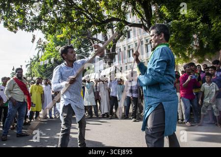 Chattogram, Bangladesch - 29. Juli 2023: Blick auf zwei Männer, die in einem heftigen Stockkampf verwickelt sind, umgeben von einer animierten Menschenmenge unter dem verfleckten Sonnenlicht, das durch üppige grüne Bäume filtert. Stockfoto