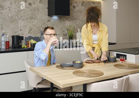 Ein Mann sitzt an einem hölzernen Esstisch und schlürft einen Drink, während eine Frau auf der Arbeitsplatte in einer stilvollen Küche eine Mahlzeit zubereitet. Die Atmosphäre ist gemütlich und Stockfoto