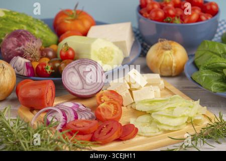 Frisches und geschnittenes Gemüse mit Feta auf einem Holzbrett, umgeben von Basilikum und Rosmarin, rustikale Küche Stockfoto