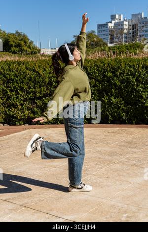 Sorglose junge Frau, die Musik mit Kopfhörern genießt und im Stadtpark frei tanzt Stockfoto