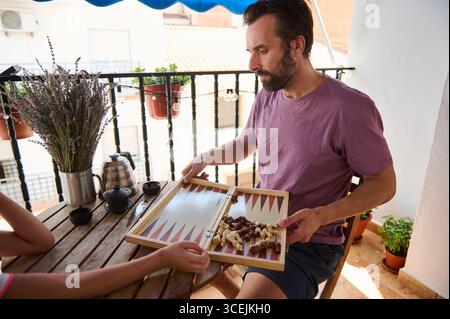 Ein Mann sitzt auf einem sonnigen Balkon, der sich mit einem Backgammon-Spiel beschäftigt. Die mit Pflanzen und Tee dekorierte ruhige Umgebung lädt zu Entspannung und Qualität ein Stockfoto