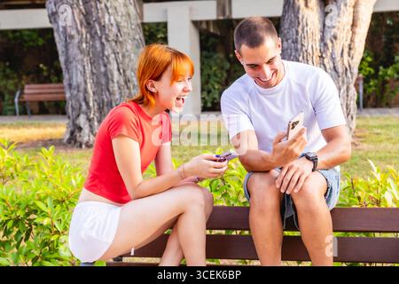 Junge Paare lachen, während sie Smartphones und Kreditkarten auf der Parkbank benutzen Stockfoto