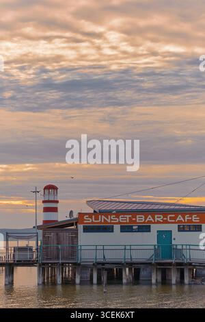 Bezauberndes Café an einem Pier am Neusiedler See, Österreich, mit atemberaubendem Blick auf den Leuchtturm Podersdorf bei Sonnenuntergang über ruhigem Wasser. Stockfoto