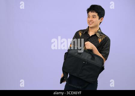 Ein Mann, der eine Aktentasche mit Batikoutfit vor violettem Hintergrund hält und zögerlich aussieht Stockfoto
