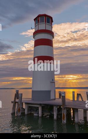 Der Leuchtturm steht hoch, während goldene Strahlen durch Wolken dringen und dramatisches Licht über den ruhigen Neusiedler See werfen. Filmisch, ruhig und atmos Stockfoto