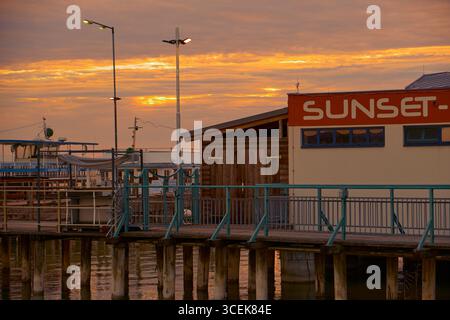 Goldener Abend über dem Pier-Café mit „Sunset“-Schild und Lampen, die ein warmes Licht auf das ruhige Wasser werfen. Stockfoto