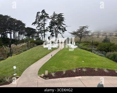 Elegante Hochzeits- und Veranstaltungseinrichtung im Freien mit weißen Tischen, Stühlen und Sonnenschirmen auf einem Rasen mit Blick auf den Pazifischen Ozean an einem nebeligen Tag Stockfoto