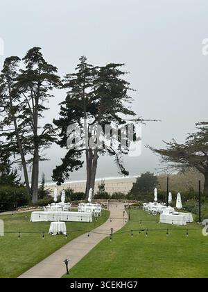 Elegante Hochzeits- und Veranstaltungseinrichtung im Freien mit weißen Tischen, Stühlen und Sonnenschirmen auf einem Rasen mit Blick auf den Pazifischen Ozean an einem nebeligen Tag Stockfoto