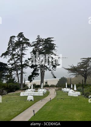 Elegante Hochzeits- und Veranstaltungseinrichtung im Freien mit weißen Tischen, Stühlen und Sonnenschirmen auf einem Rasen mit Blick auf den Pazifischen Ozean an einem nebeligen Tag Stockfoto