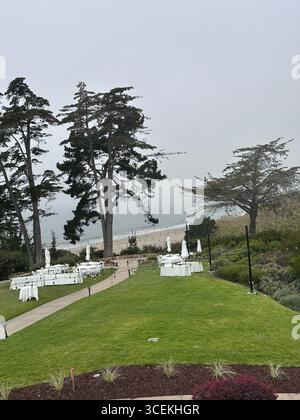 Elegante Hochzeits- und Veranstaltungseinrichtung im Freien mit weißen Tischen, Stühlen und Sonnenschirmen auf einem Rasen mit Blick auf den Pazifischen Ozean an einem nebeligen Tag Stockfoto