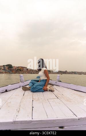 Junge Frau, die auf einem Holzboot auf dem Ganges-Fluss sitzt, die Stadtansicht von Varanasi mit bedecktem Himmel im Hintergrund bewundern und authentische Ind. Erleben Stockfoto