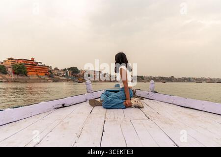 Junge Frau, die auf einem Holzboot sitzt und die atemberaubende Stadtlandschaft von Varanasi vom ruhigen Wasser des Ganges, eines der Hinduismus, genießt Stockfoto