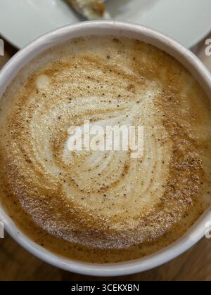 Diese Abbildung zeigt eine Ansicht von oben nach unten eines Cappuccinos, der in einem weißen Einwegbecher serviert wird. Der Schaumstoff auf der Oberseite zeigt ein klassisches, blattartiges Latte Art-Design Stockfoto