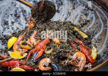 Schwarze Paella mit Meeresfrüchten. Traditionelle schwarze Paella mit Garnelen, Muscheln und Calamari, serviert mit Zitronenscheiben in einer großen Pfanne. Konzept des Spanischen Stockfoto