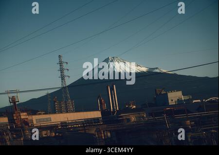 Schneebedeckter Fuji-Berg hinter Stromleitungen und Fabriken, von einem Zugfenster aus gesehen Stockfoto