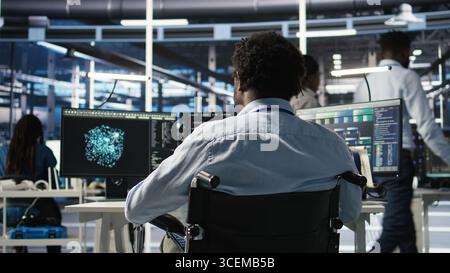 Der Programmierer im Serverraum mit Querschnittslähmung interpretiert KI-generierte Datenanalysediagramme. Mann im Rollstuhl mit künstlicher Intelligenz, das prädiktive Systemmetriken im Rechenzentrum zeigt, Kamera A Stockfoto