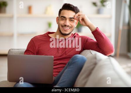 Positiver arabischer Kerl, der sich auf der Couch mit Laptop zurücklehnen kann Stockfoto