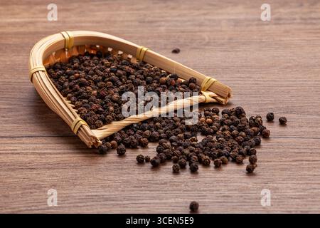 Schwarze Pfefferkörner im Bambuskorb auf Holztisch - kulinarischer Hintergrund Stockfoto
