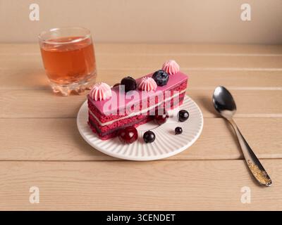 Ein Stück Kuchen mit rosa Zuckerguss, verziert mit Heidelbeeren und Kirschen, serviert auf einem weißen Teller. Begleitet von einem Glas Orangengetränk und Stockfoto