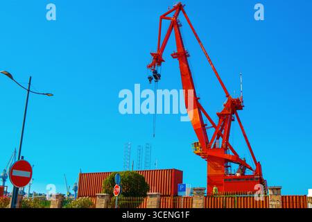 Markanter roter Industriekran ragt über Schiffscontainern unter einem hellblauen Himmel und hebt die Energie eines geschäftigen Hafens hervor. Kräftige Farben und starke Farben Stockfoto