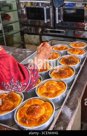 Älterer Bäcker veredelt goldene Käsebrotlaibe in runden Metallpfannen in einer rustikalen Bäckerei. Stockfoto